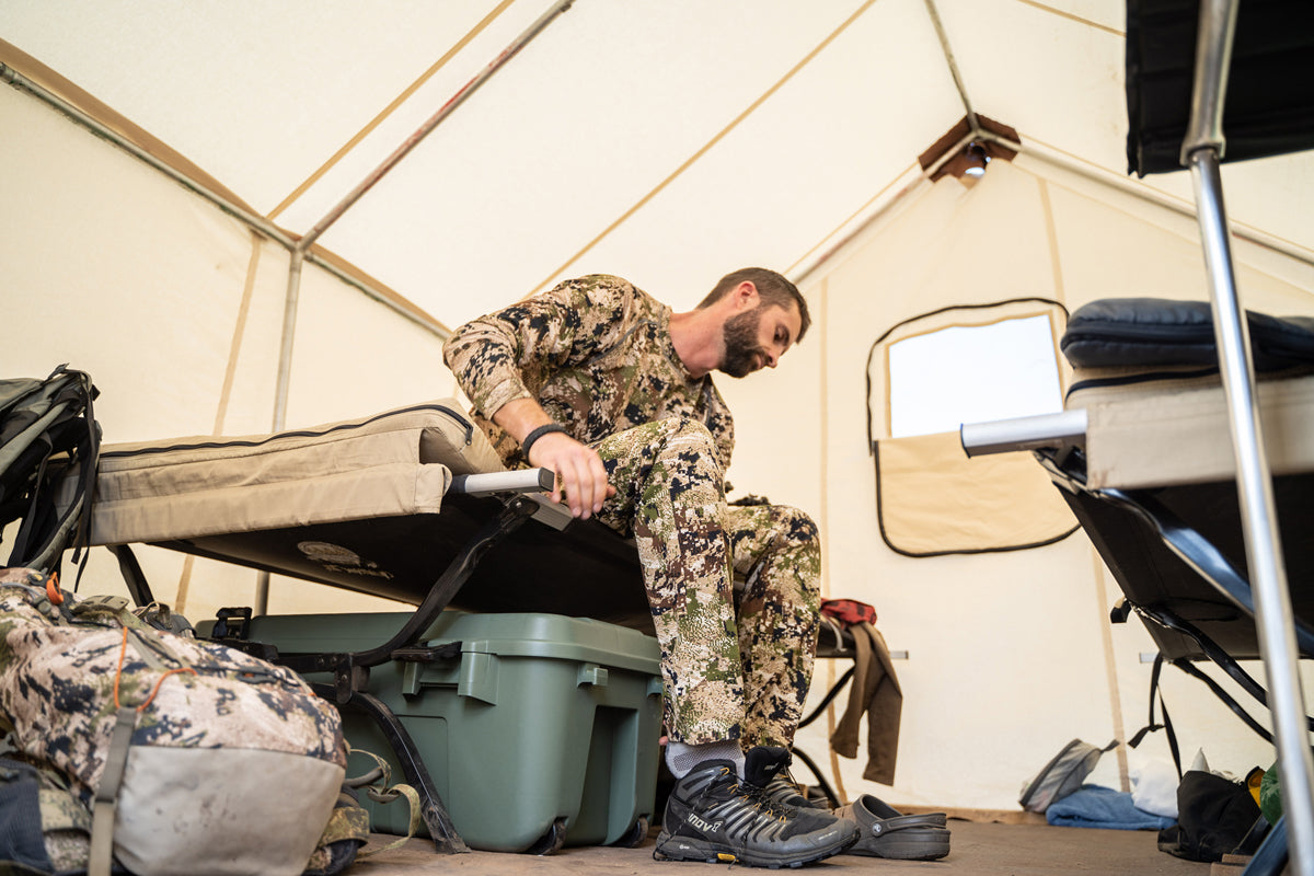 A man in camo gear sits on a cot inside a tent, putting on his shoes. Camping gear and a backpack are on the floor nearby. The tent has a white interior and simple furnishings.