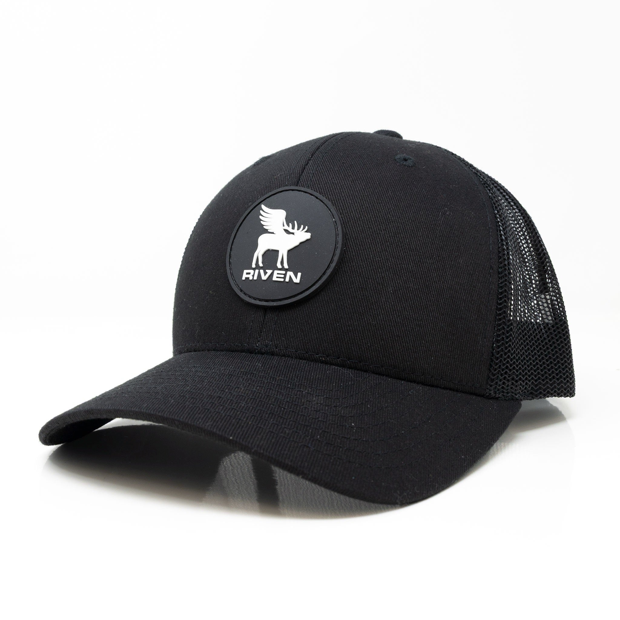 The Riven Flying Elk Hat is a black trucker cap with a curved brim, mesh back, and adjustable snap closure. A round patch featuring a white elk and "RIVEN" decorates the front. One size fits most. Shown on a plain white background.