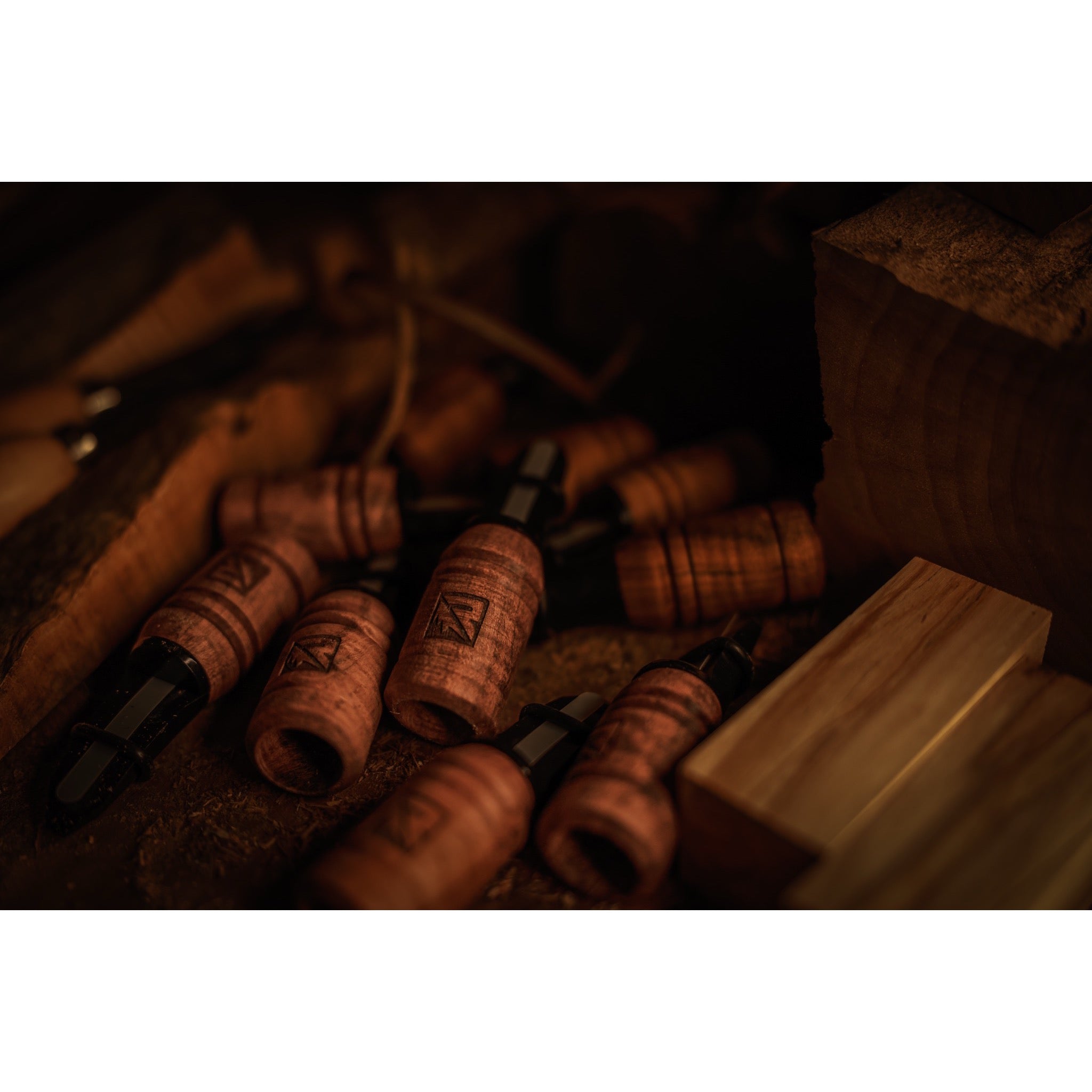 Riven Menace-Maple wooden duck calls with dark mouthpieces are scattered on a wooden surface among wood pieces in warm, low lighting.