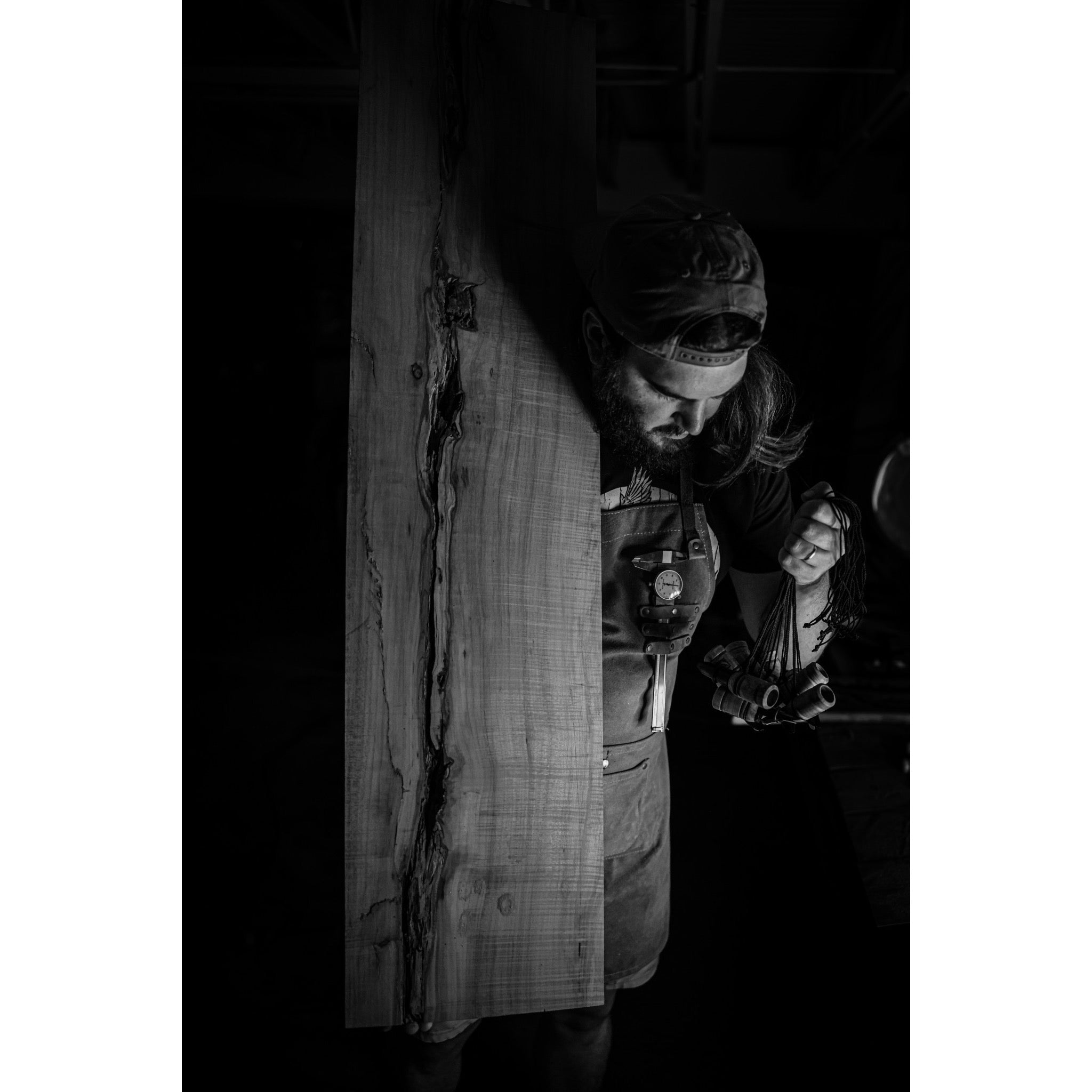 A man in an apron and backward cap carries a Menace-Maple wooden plank by Riven on his shoulder, tools in hand. The black-and-white image features dramatic lighting and deep shadows.