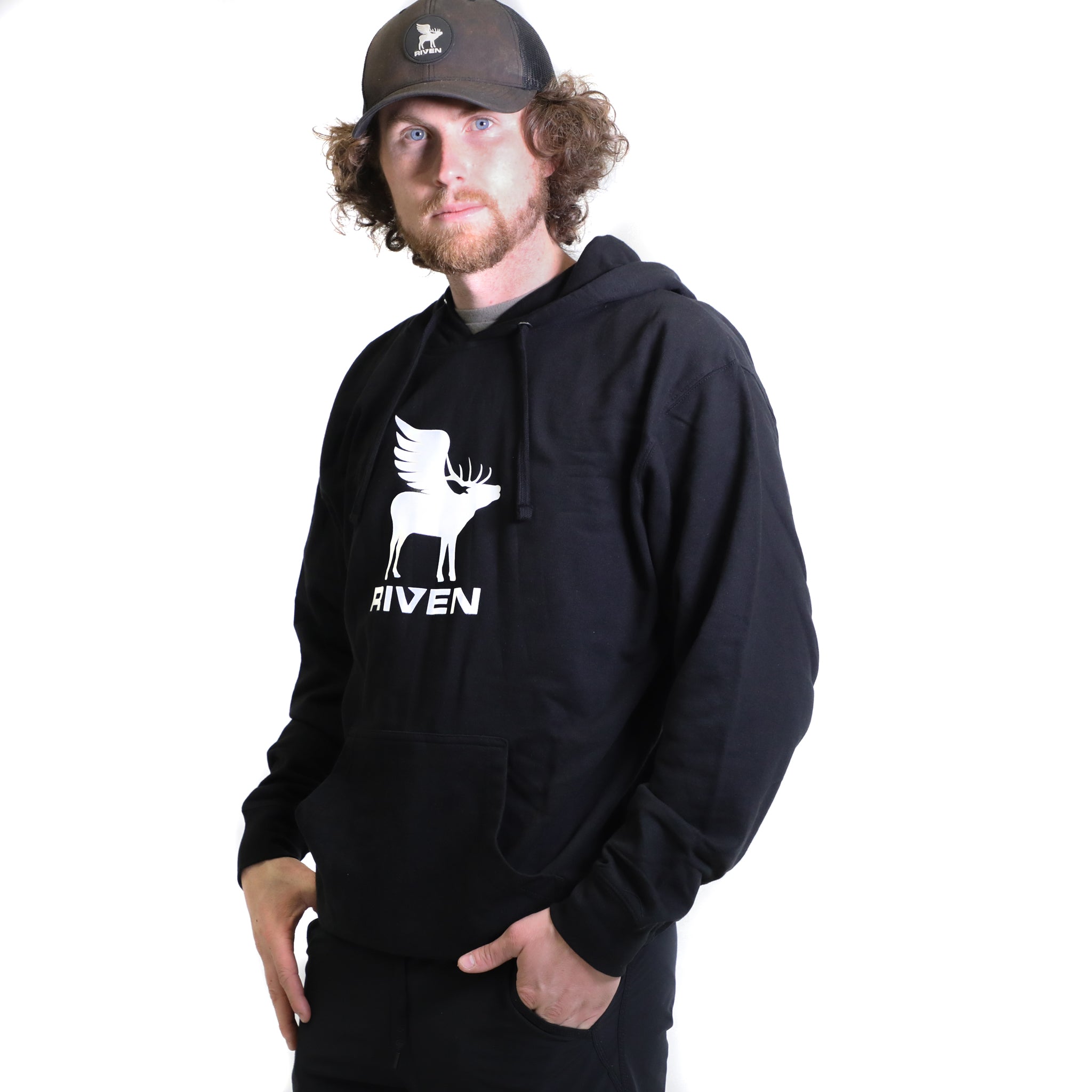 A man with curly hair and a beard wears the Riven Flying Elk Hoodie, which is black with a white winged elk graphic and "RIVEN" text. He also sports a black cap with a white animal logo, standing before a plain white background.