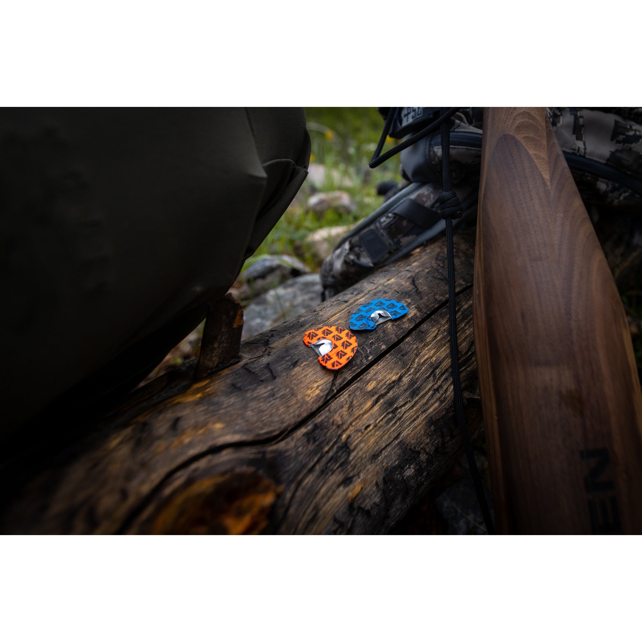 Two colorful heart-shaped objects, one orange and one blue, rest on a weathered log outdoors. A Riven Cambium Elk Bugle Tube and a backpack are nearby, with grass and rocks in the background.