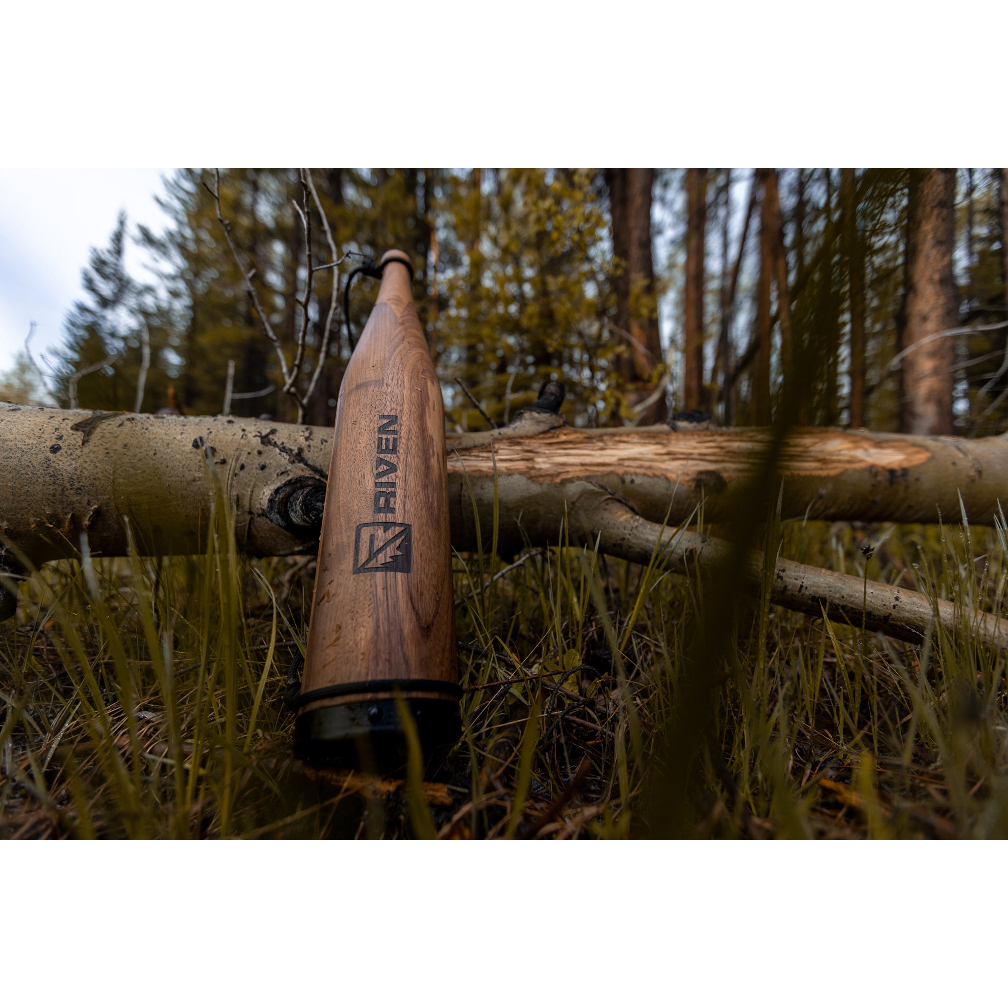 A Cambium Elk Bugle Tube by Riven lies on grass in a forest, leaning against a fallen tree, surrounded by trees and greenery.
