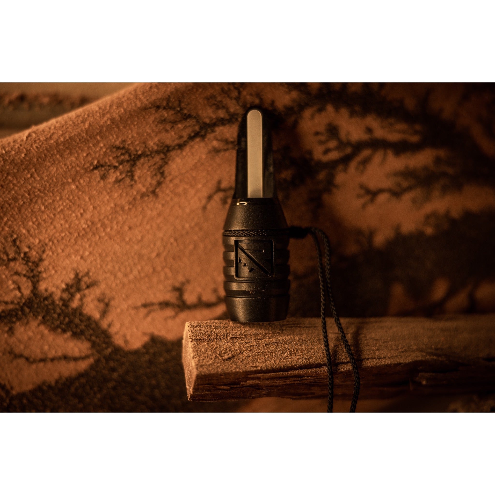 The Riven Menace black duck call with lanyard rests on wood, set against a warm camo background. Its bite and blow design offers versatility for waterfowl hunts.