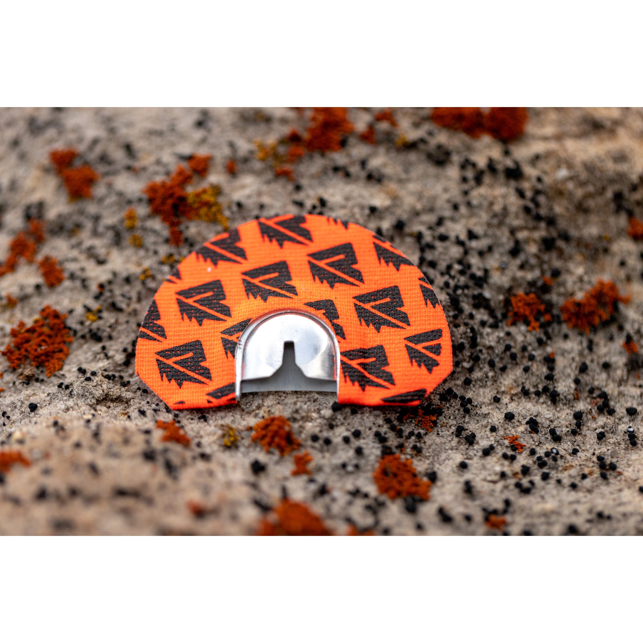 The Riven Tremor, an orange and black turkey hunting diaphragm call with a metal reed, rests on a rocky surface with matching lichen, resembling an elk call for mimicking cow elk sounds in the wild.