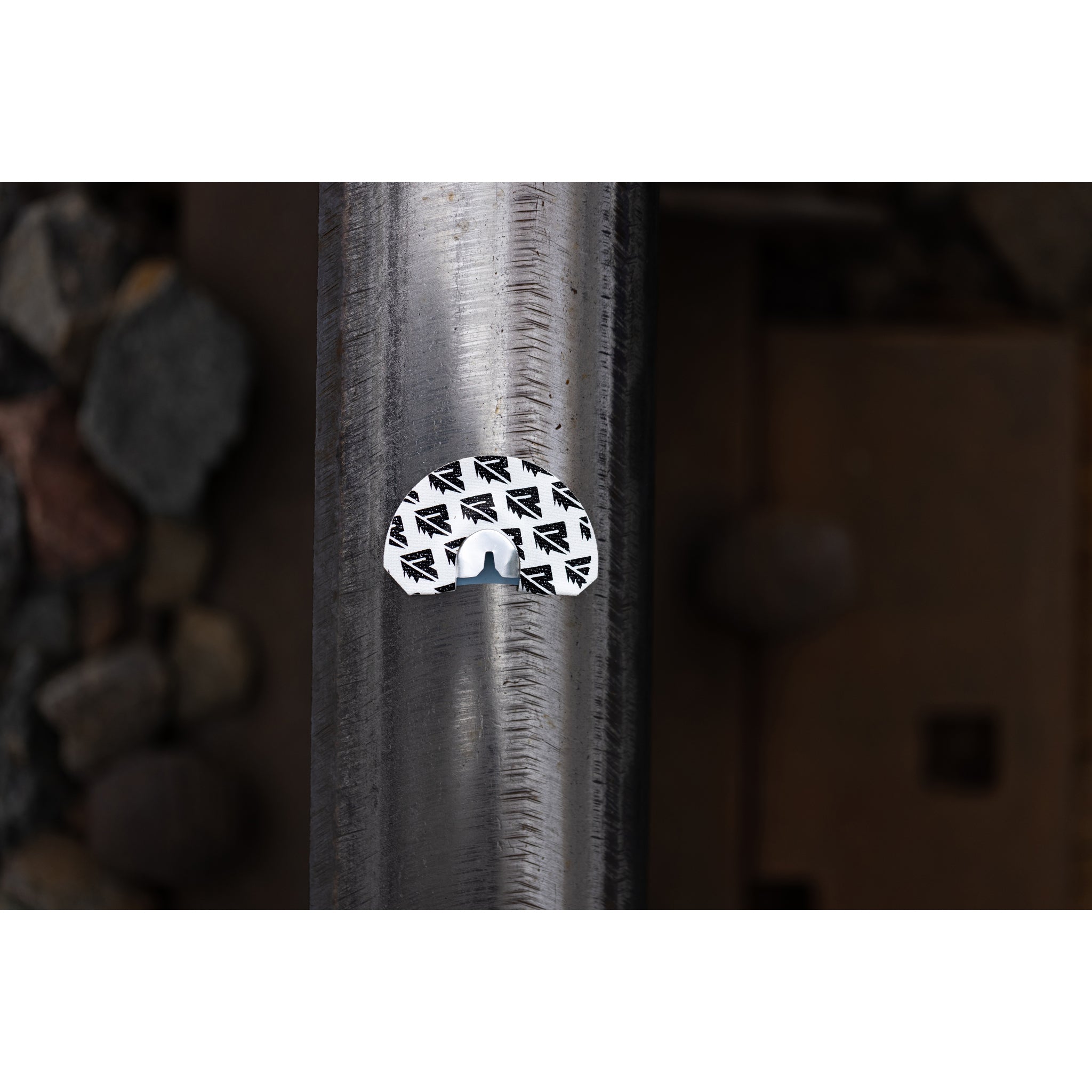 A white, semi-circular Riven Rosie sticker with a black repeating pattern—shaped like an elk mouth call—is attached to a metal rail, with rocks and blurred brown in the background.