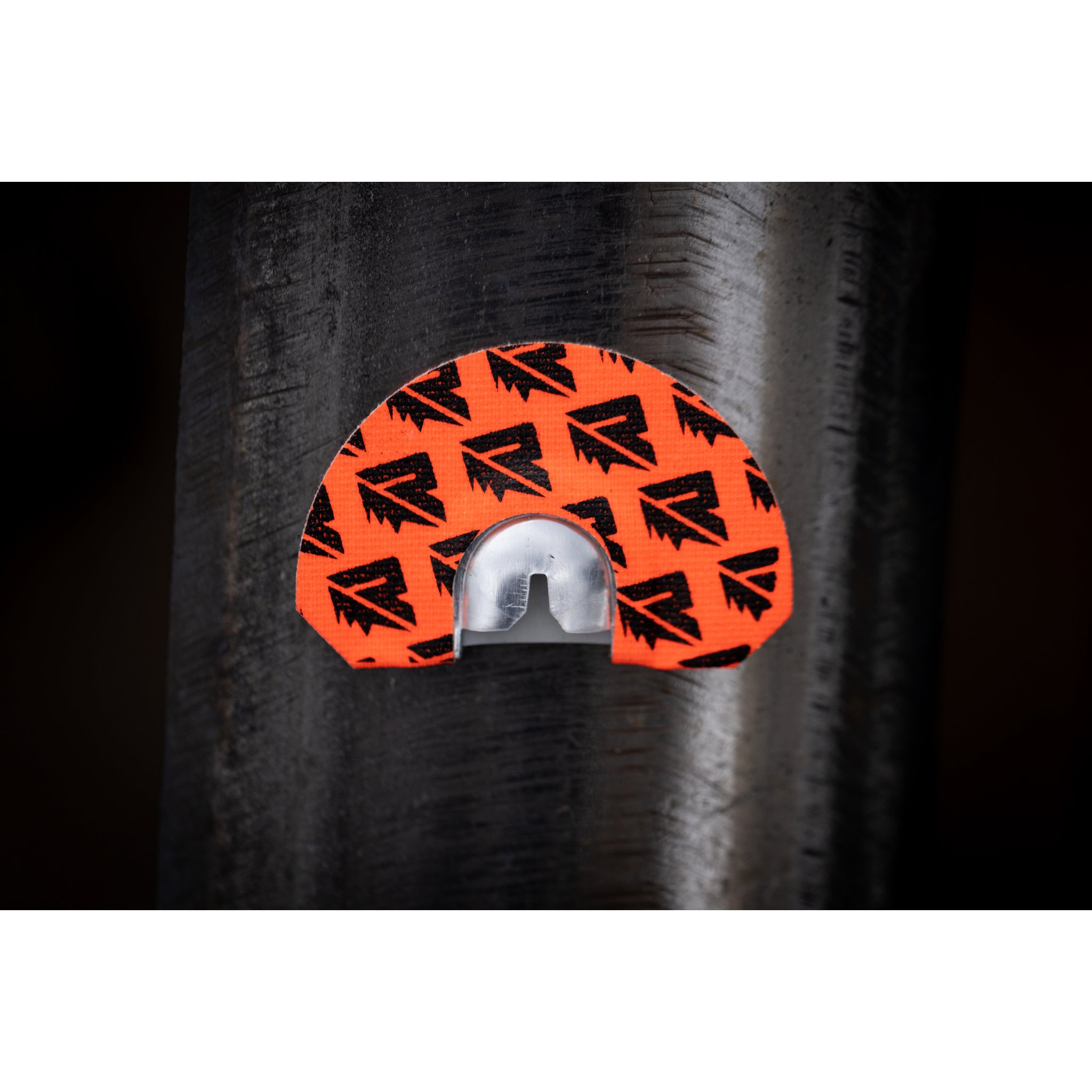 A close-up of the Riven Tremor: an orange and black sticker with arrow patterns and a metallic sensor, attached to a dark cylindrical surface—evoking the sharp detail of cow elk calls in the wild.
