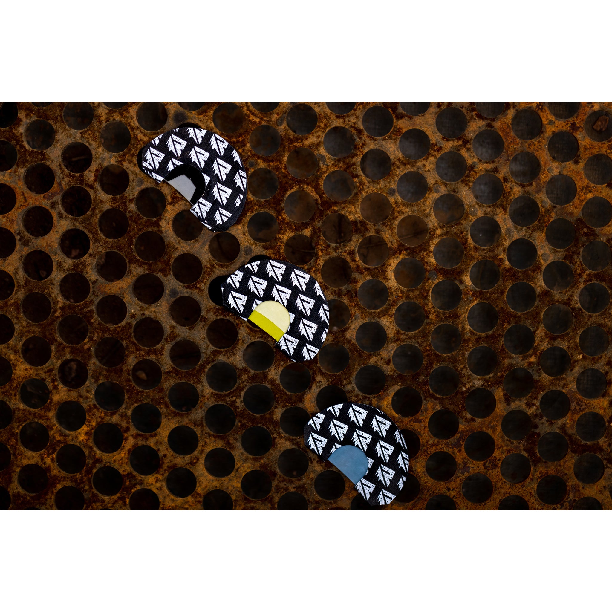Three Riven Origin Series elk mouth calls with white arrow patterns—each featuring unique color accents in yellow, blue, or solid black—are arranged diagonally on a rusty, perforated metal surface.