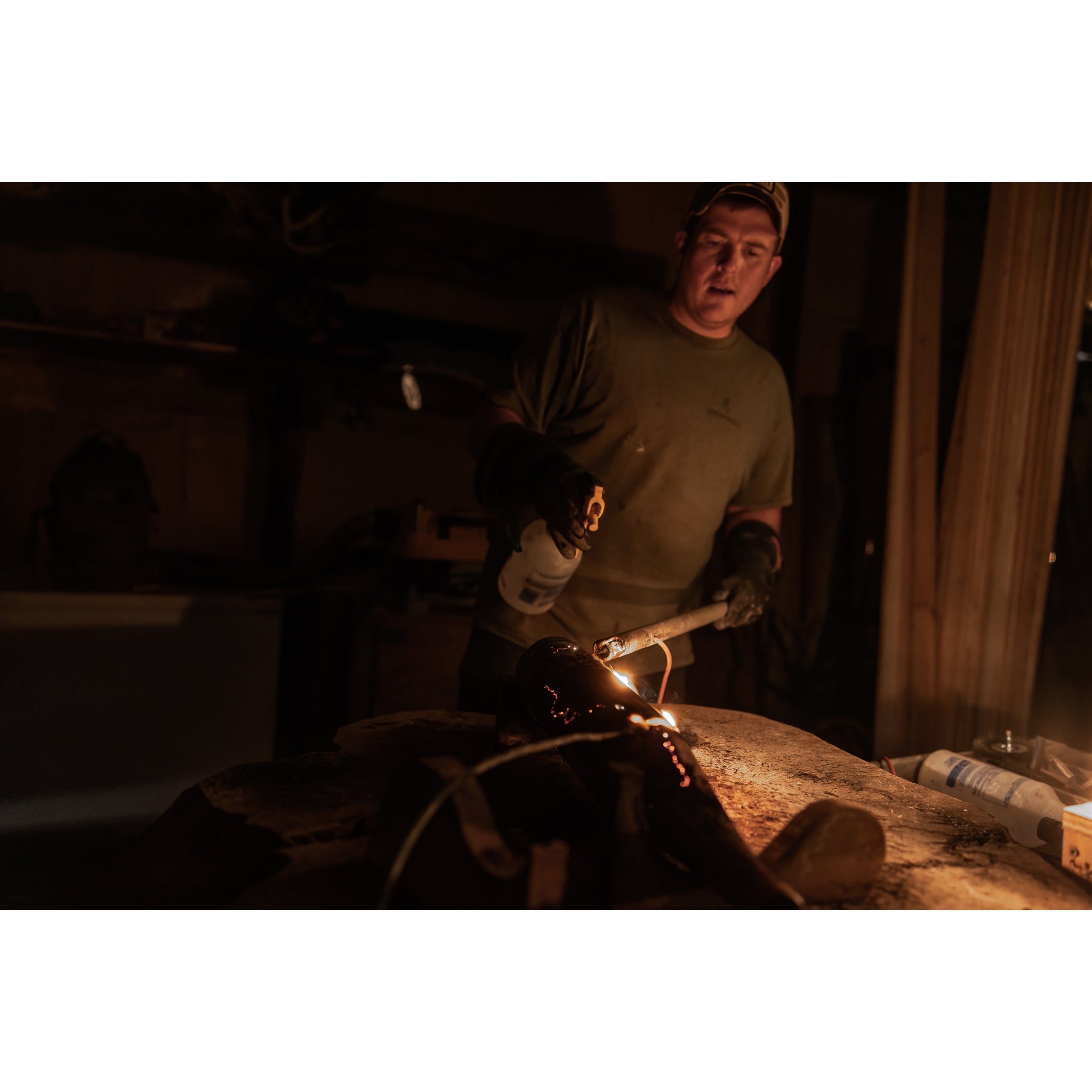 Wearing gloves and a cap, a person uses a blowtorch in a dim workshop, sparks flying as they craft the Riven Rough Cut Cambium - Mystery Storm Breaker with its unique hand-crafted finish and limited edition colors gleaming.