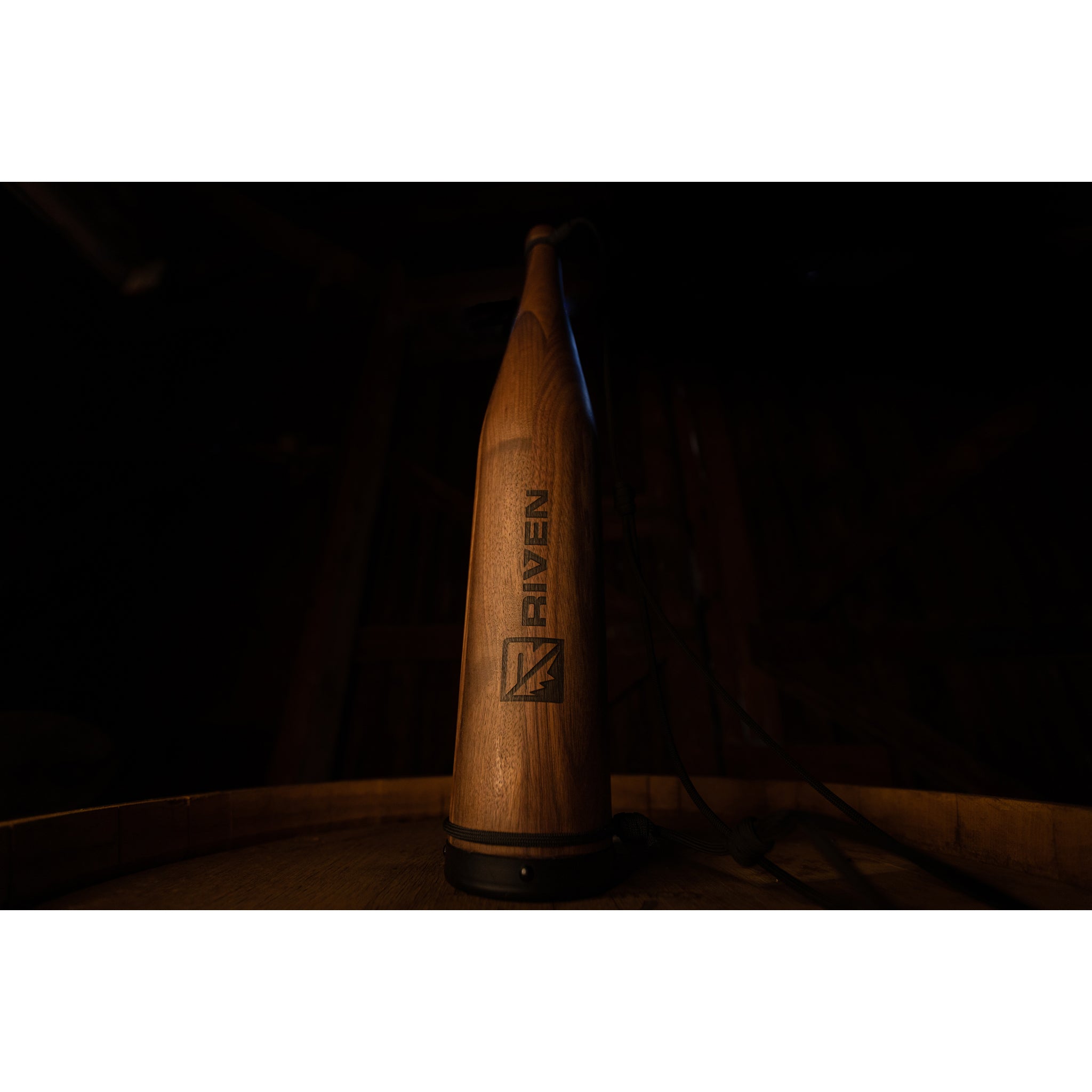 A tall Cambium Elk Bugle Tube by Riven, crafted from natural tonewood and etched with the Riven logo, stands upright on a barrel against a dark, blurred background.