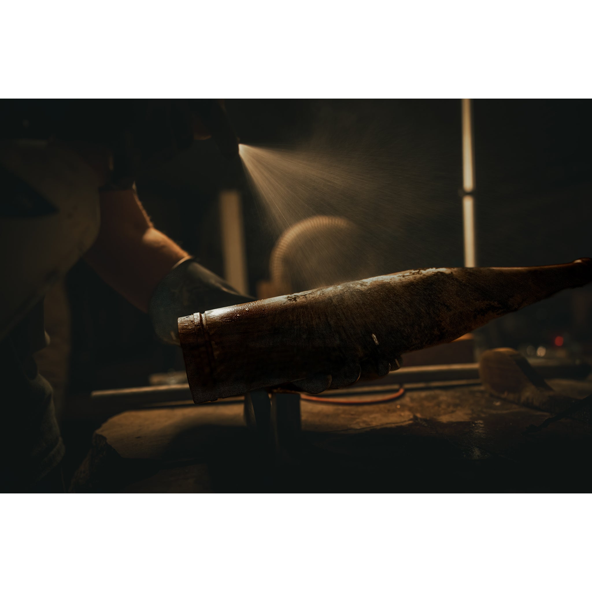Wearing gloves, a person sprays Riven's Rough Cut Cambium - Mystery Storm Breaker onto a weathered metal object in a dim workshop, dramatic lighting emphasizing the mist and hand-crafted finish.