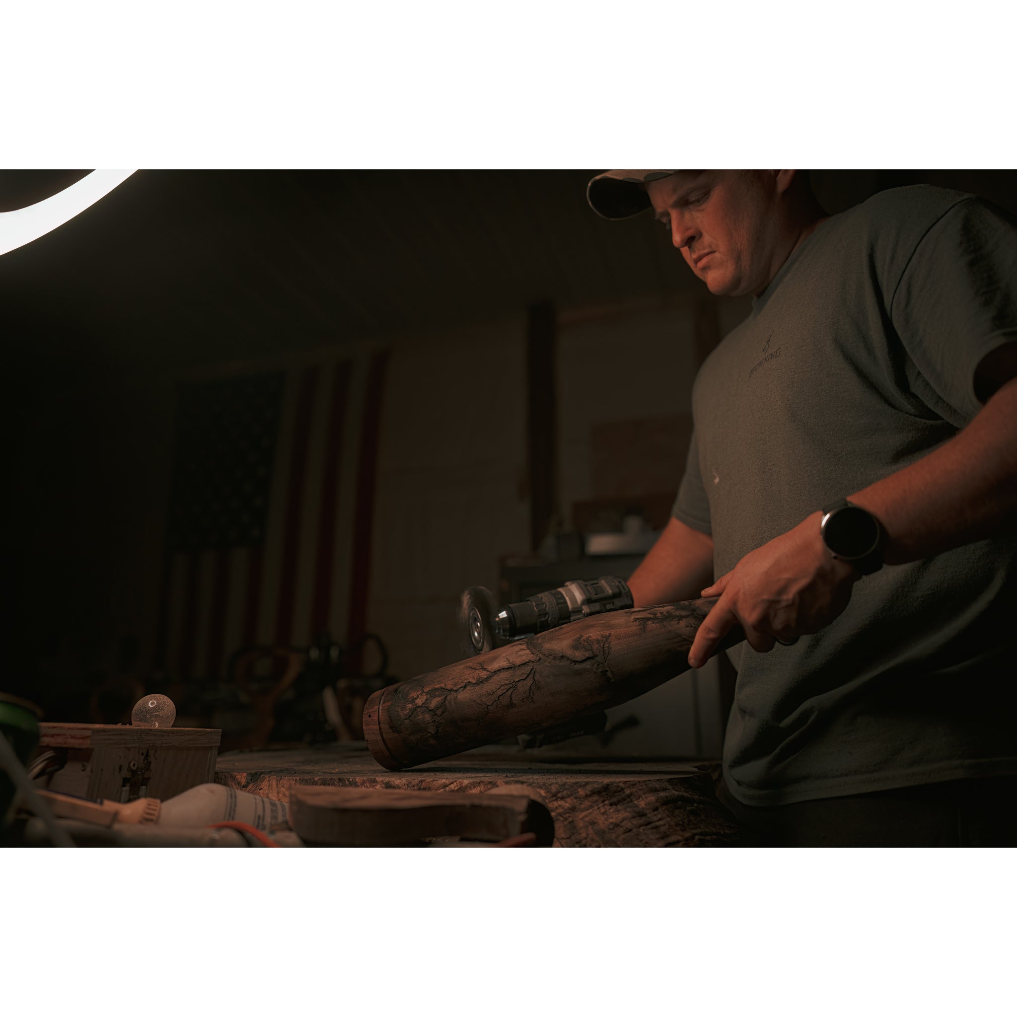 In a dimly lit workshop with an American flag, a person in a cap uses a power tool on wood with the unique hand-crafted finish of the Riven Rough Cut Cambium - Mystery Storm Breaker.