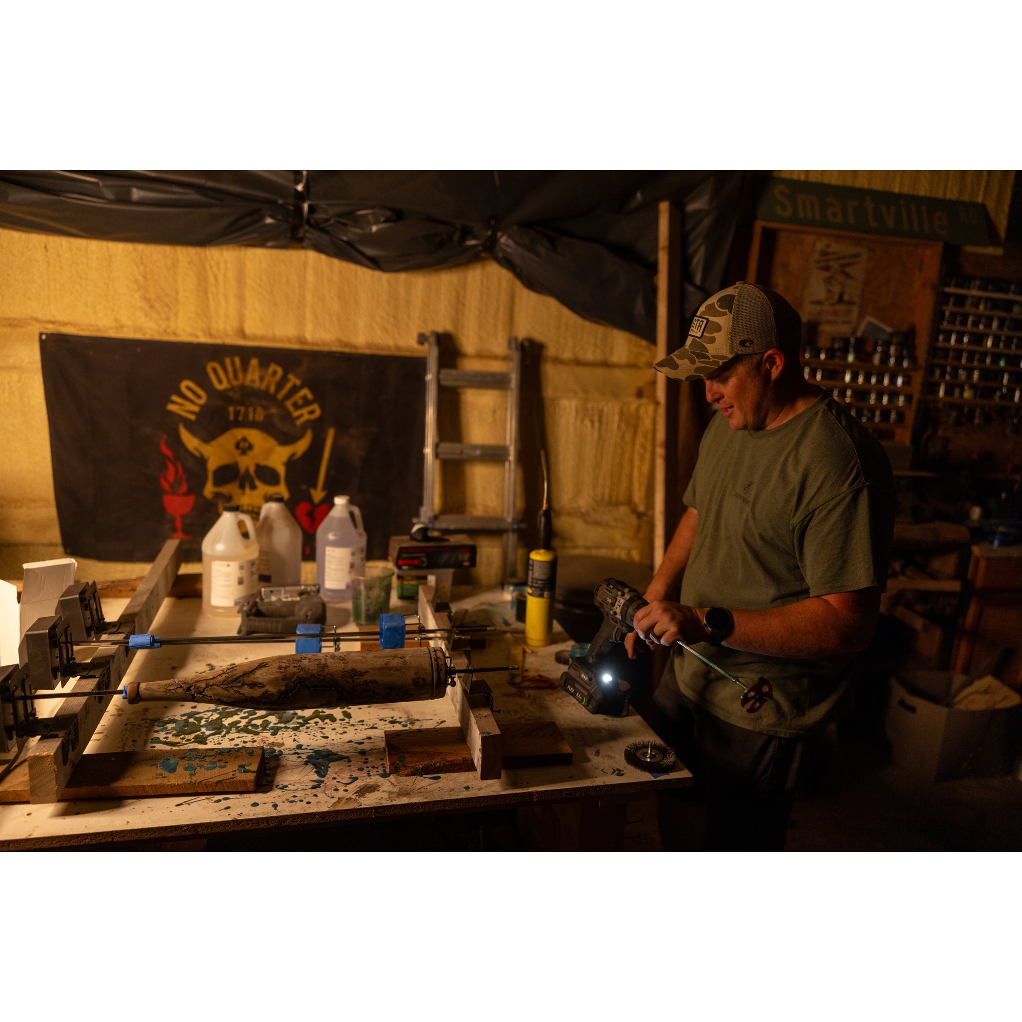 A man in a cap works in a dimly lit workshop, his table cluttered with tools and materials. Behind him hangs a No Quarter pirate flag as he crafts the Riven Rough Cut Cambium - Mystery Storm Breaker with a unique hand-crafted finish.