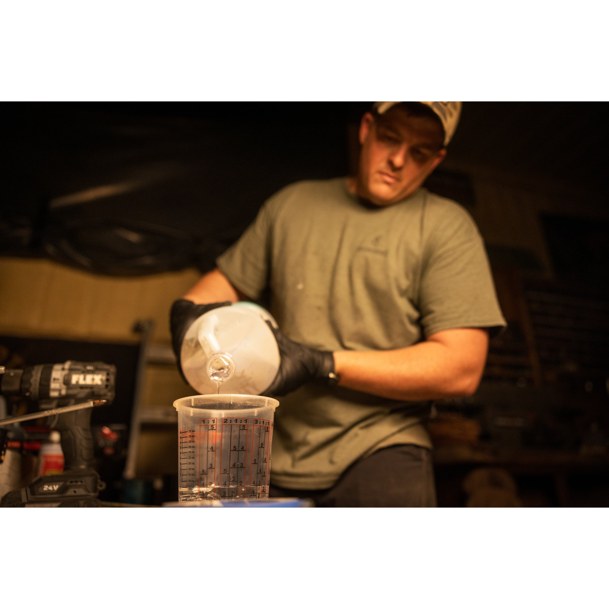 A person in a green shirt and black gloves pours liquid from a jug into a clear measuring cup on a workbench, preparing materials for the Riven Rough Cut Cambium - Mystery Storm Breaker with its unique hand-crafted finish.