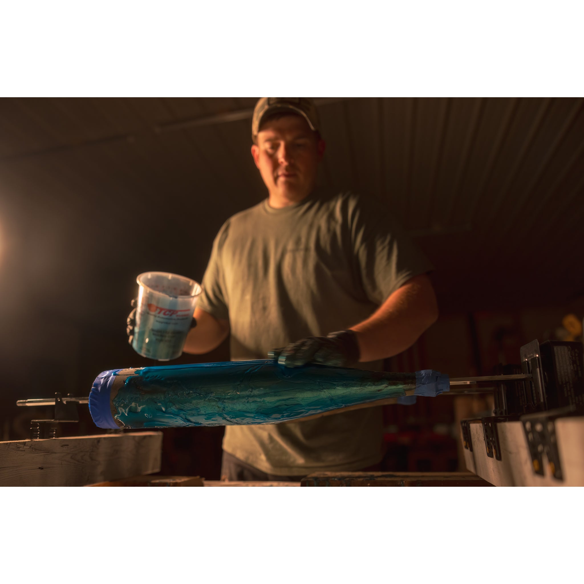 In a dim workshop, a person in a cap and gloves pours liquid onto a blue-wrapped cylinder, expertly hand-finishing the Riven Rough Cut Cambium - Mystery Storm Breaker for an exclusive touch.