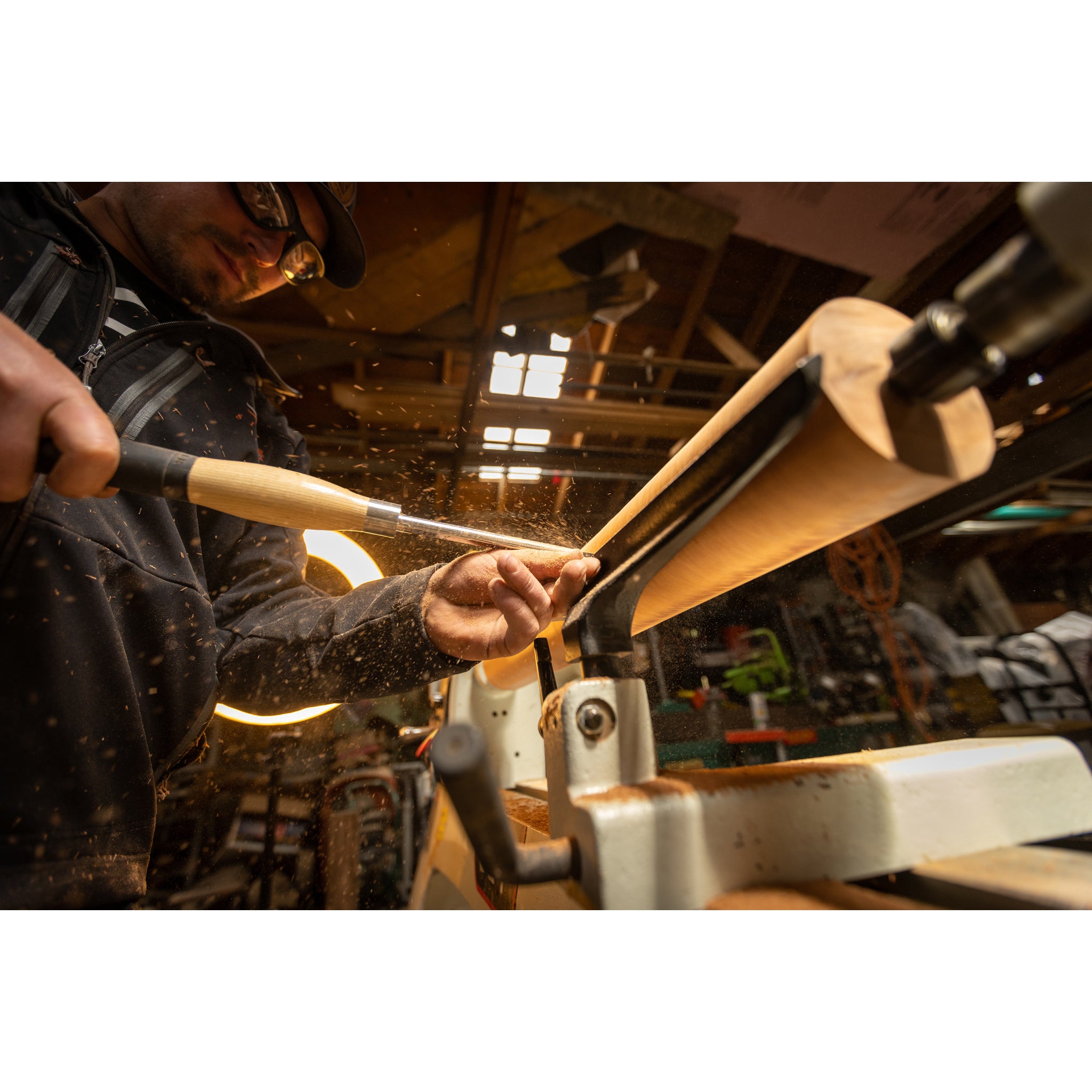 A person shapes a Cambium Elk Bugle Tube by Riven in a workshop, surrounded by wood shavings and tools, as warm light highlights the natural tonewood.
