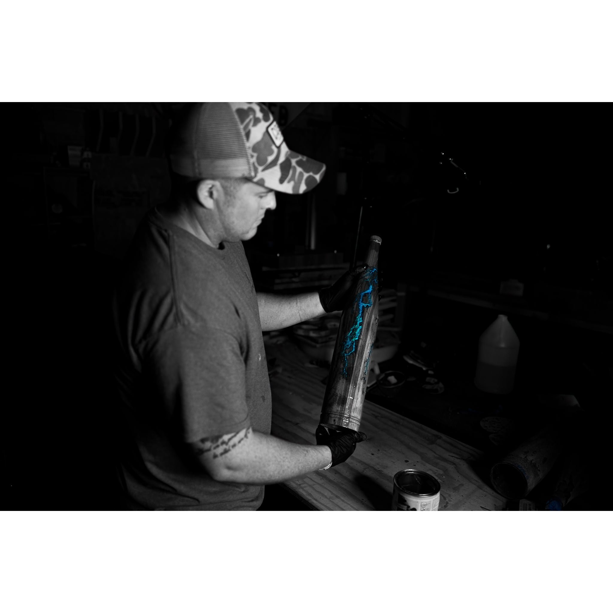 A man in a camo hat and gloves holds Riven’s Rough Cut Cambium - Mystery Storm Breaker, a limited edition cylindrical object with a blue lightning design, vividly colored against the black-and-white backdrop of a dim workshop.