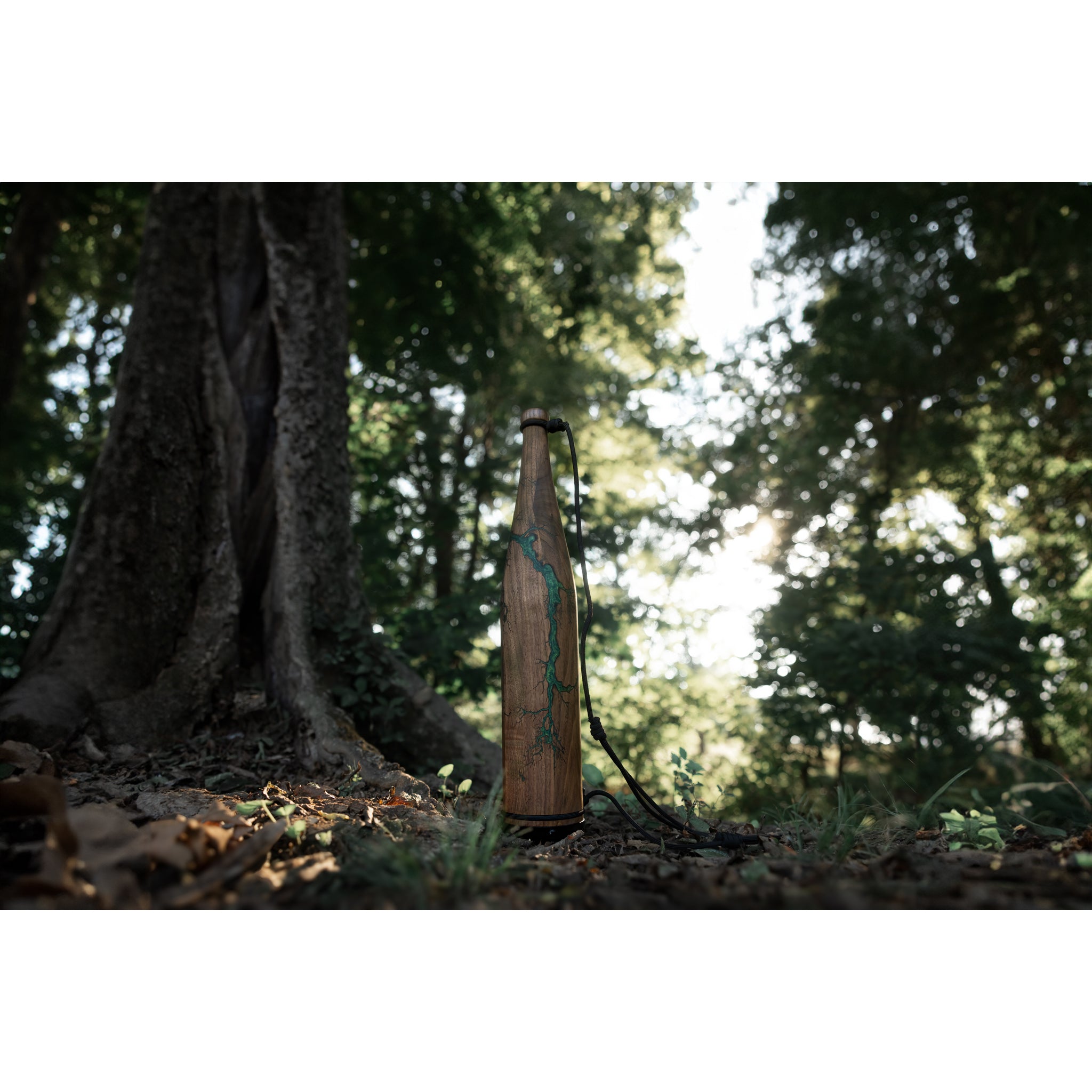 The Riven Rough Cut Cambium - Mystery Storm Breaker, a wooden water bottle with a green design and black strap, stands upright by a tree on the forest floor, highlighting its unique hand-crafted finish amid greenery and sunlight.