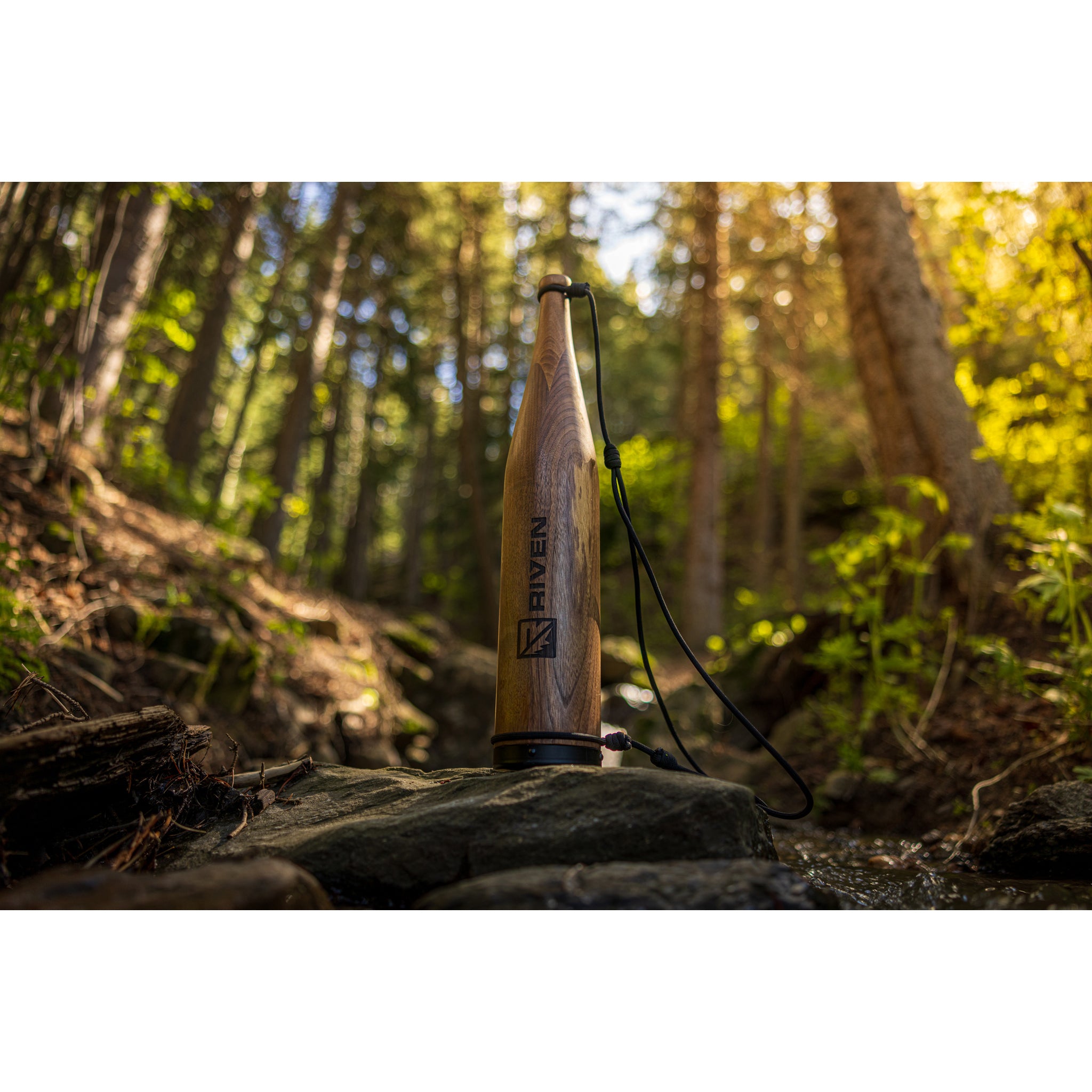 A Cambium Elk Bugle Tube by Riven rests on a rock in a sunlit forest, surrounded by lush greenery and tall trees. Sunlight streams through the branches, creating a tranquil, natural setting.