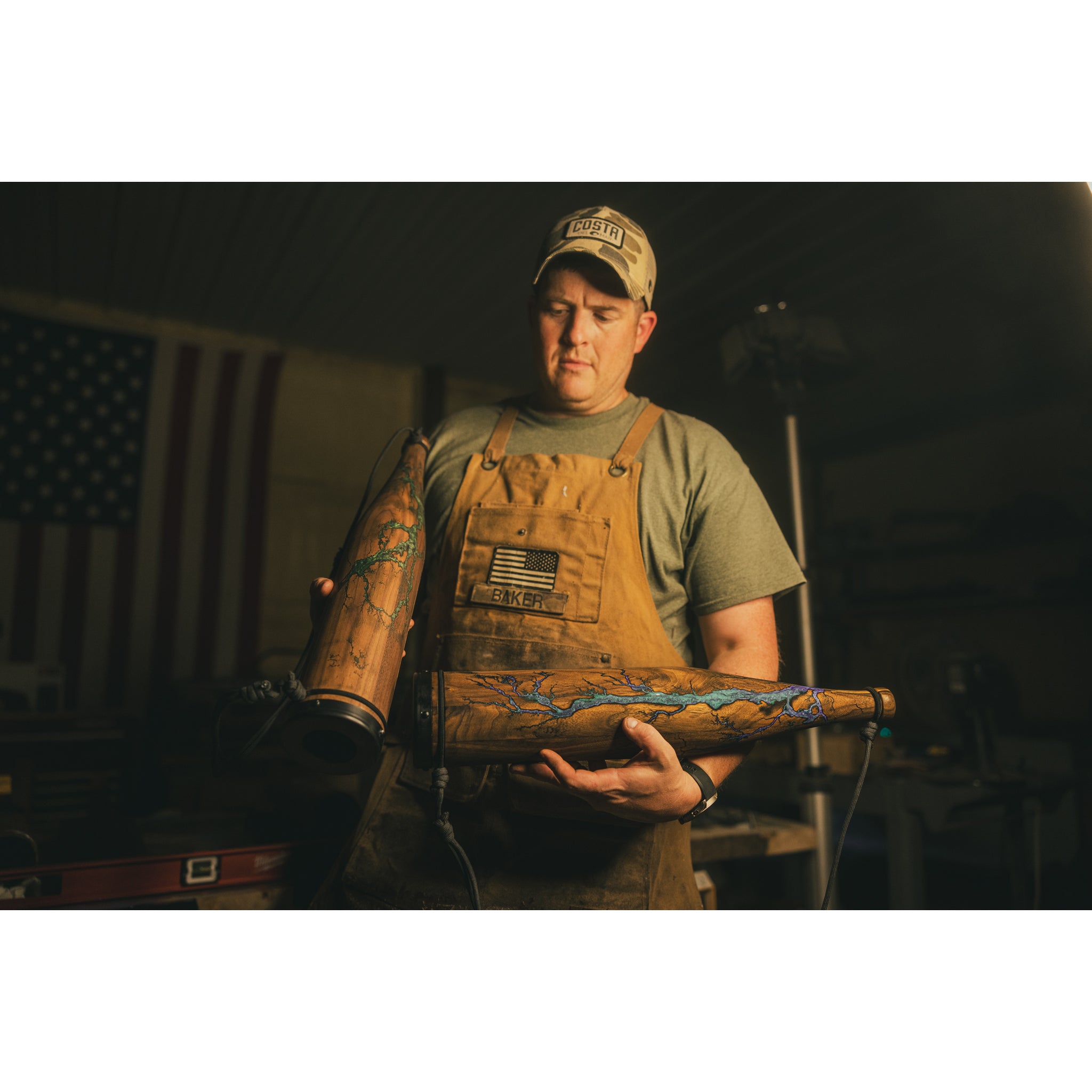 In a warmly lit workshop, a person in a brown apron and cap holds two Riven Rough Cut Cambium - Mystery Storm Breaker exhaust pipes with hand-crafted finishes. An American flag hangs in the background, highlighting these special edition creations.