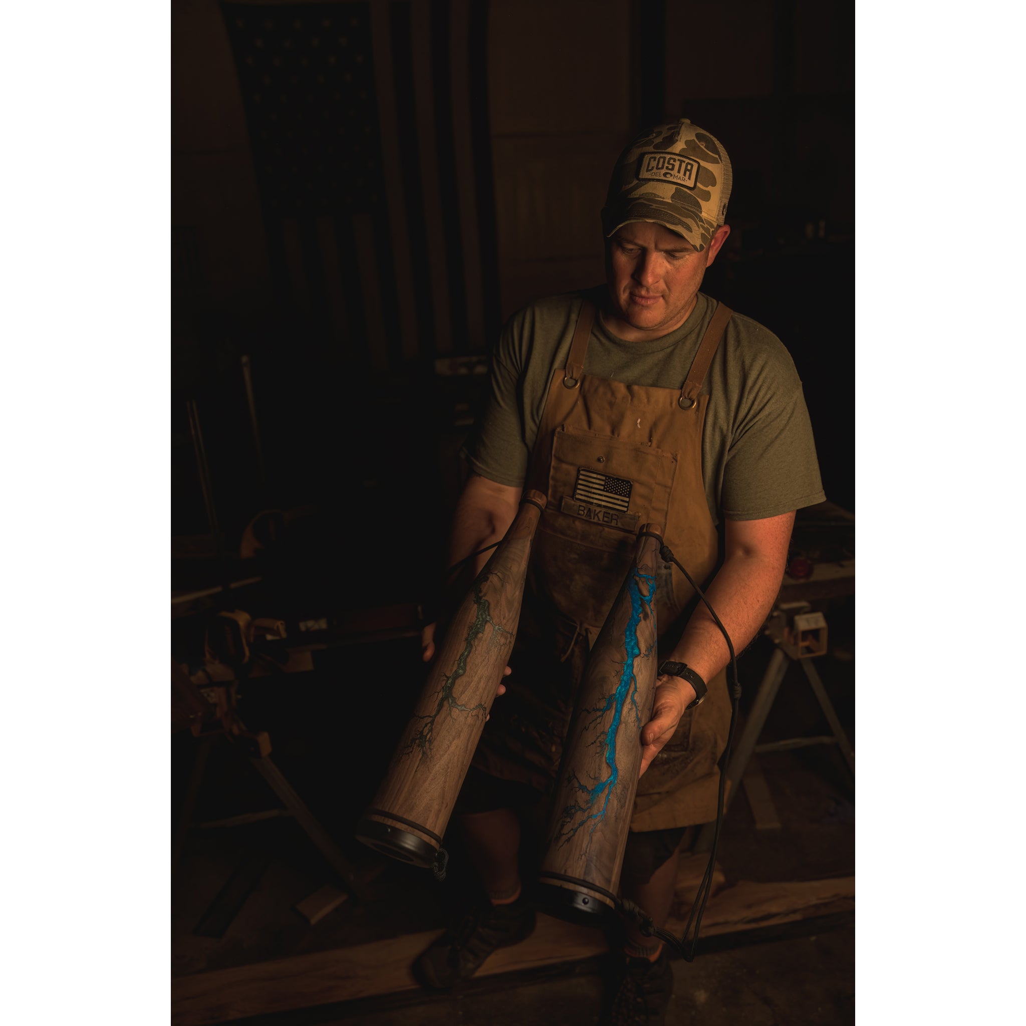 A person in a cap, glasses, and brown apron indoors holds two Riven Rough Cut Cambium - Mystery Storm Breakers, featuring unique handcrafted finishes with blue accents; an American flag is visible in the dim background.