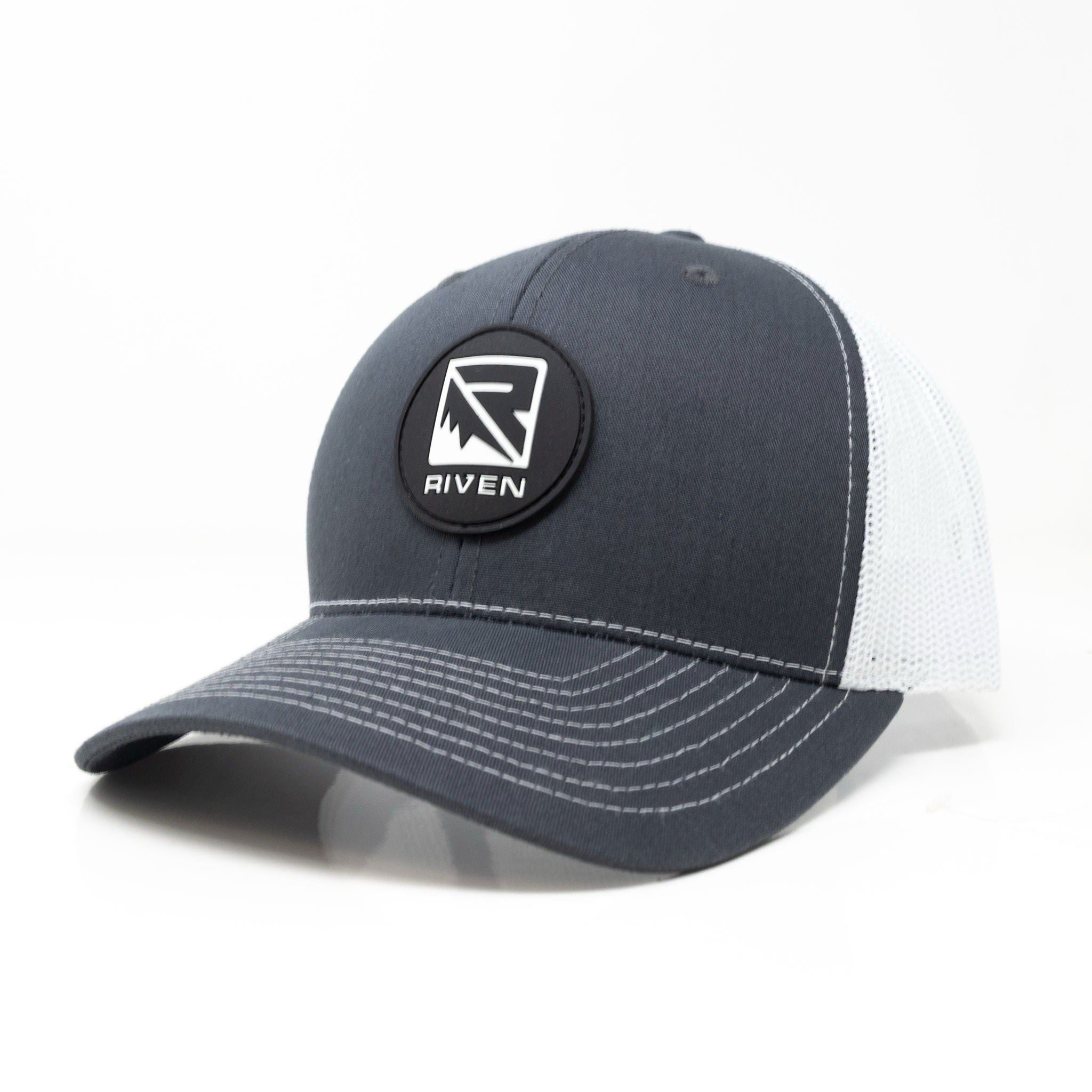 The Riven Patch Hat is a dark gray and white trucker hat featuring white stitching, a black patch with the Riven logo, and a one-size-fits-most snapback mesh design for comfort.